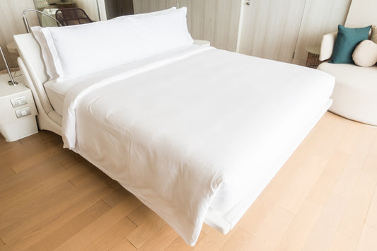 A double bed made with white linen, including a fitted sheet, duvet cover, and pillowcases, in a room with wooden flooring and a couch in the background.